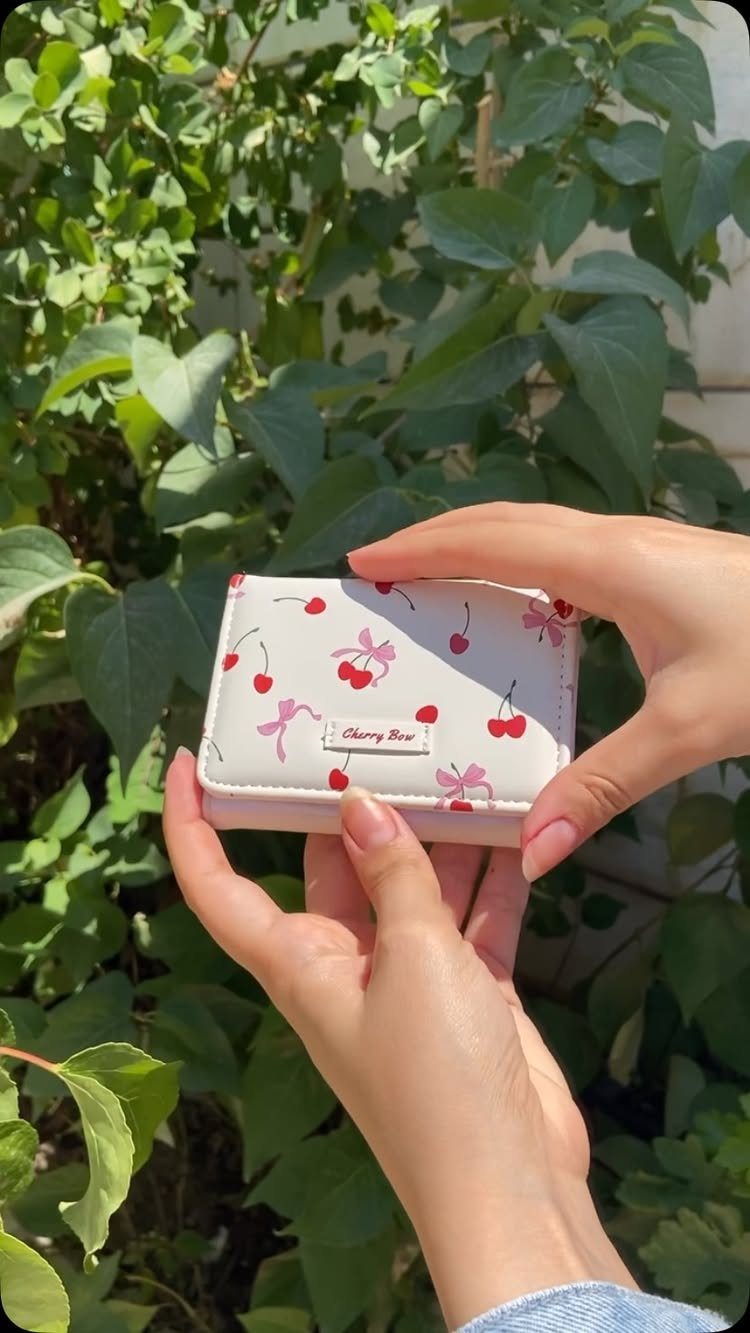 Small white wallet with red cherry patterns held against a background of green leaves.