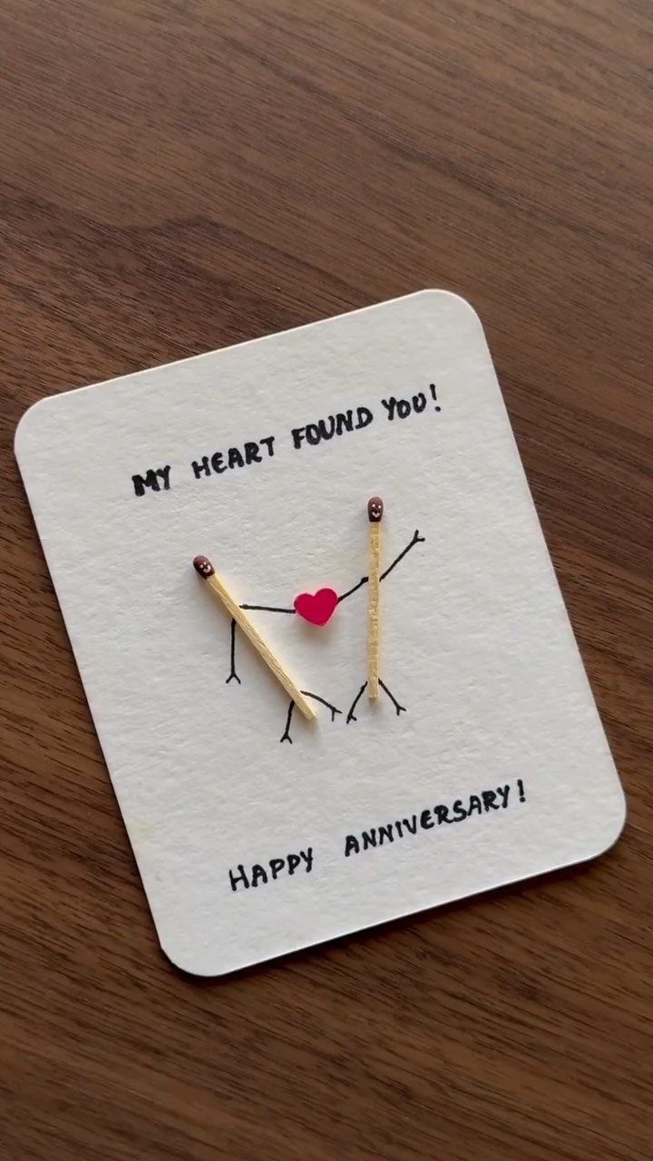 Two matchsticks as figures holding hands with a red heart on a card.
