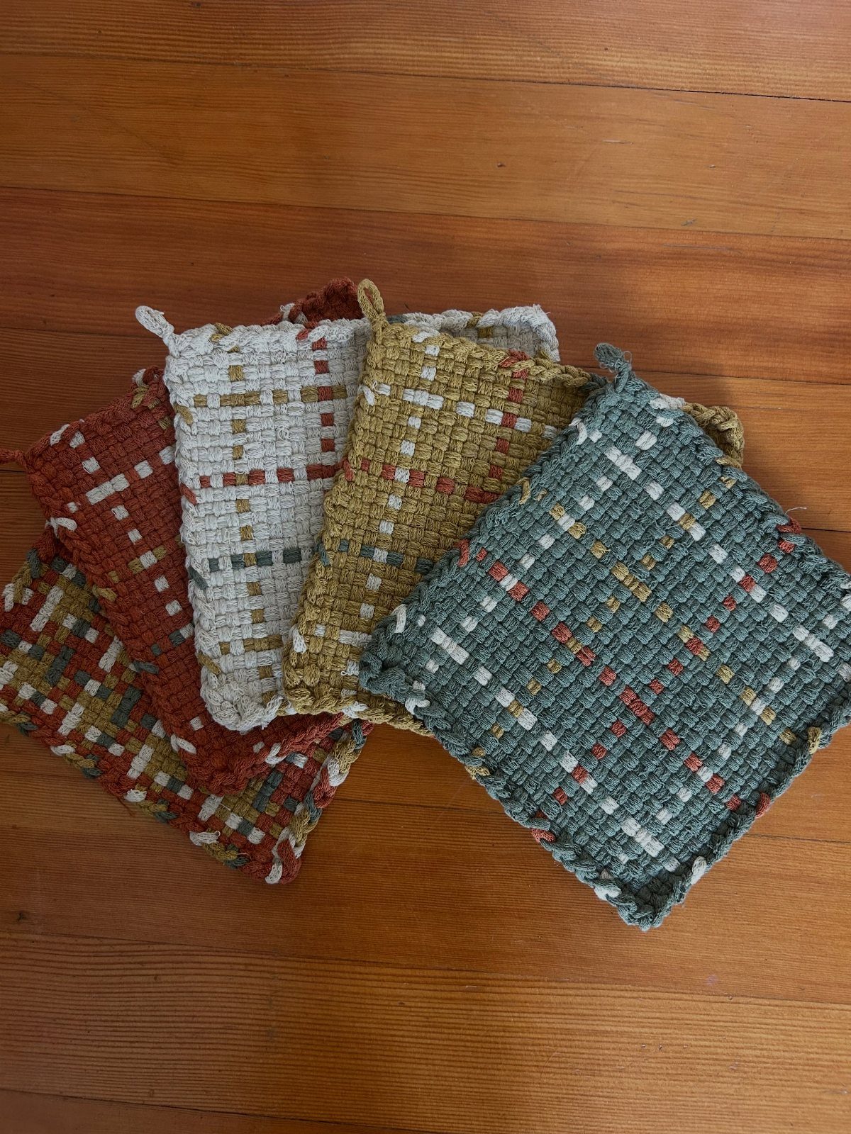 Five colorful woven fabric potholders arranged on a brown wooden floor.