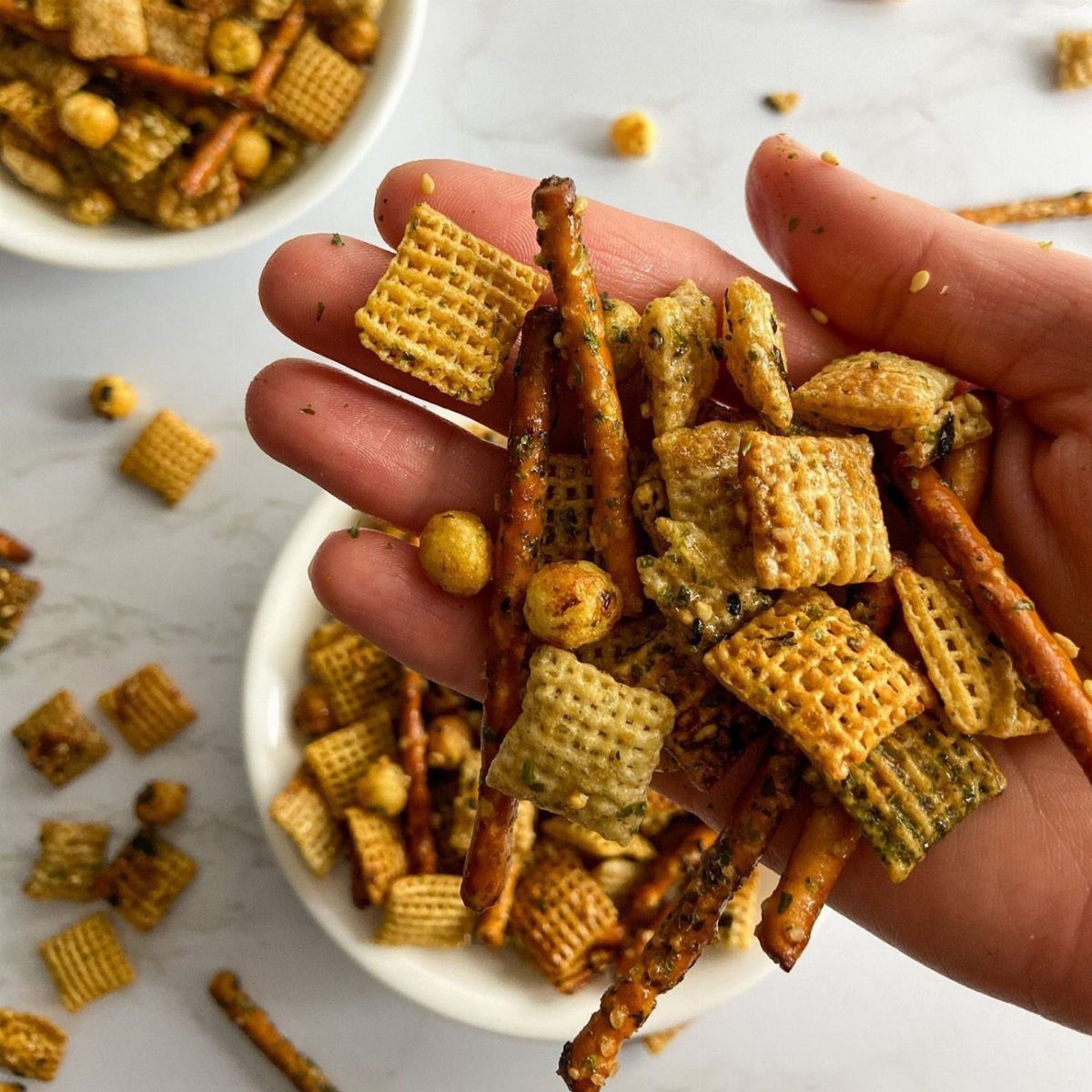 Savory snack mix with cereal and pretzels held in a person's open hand.