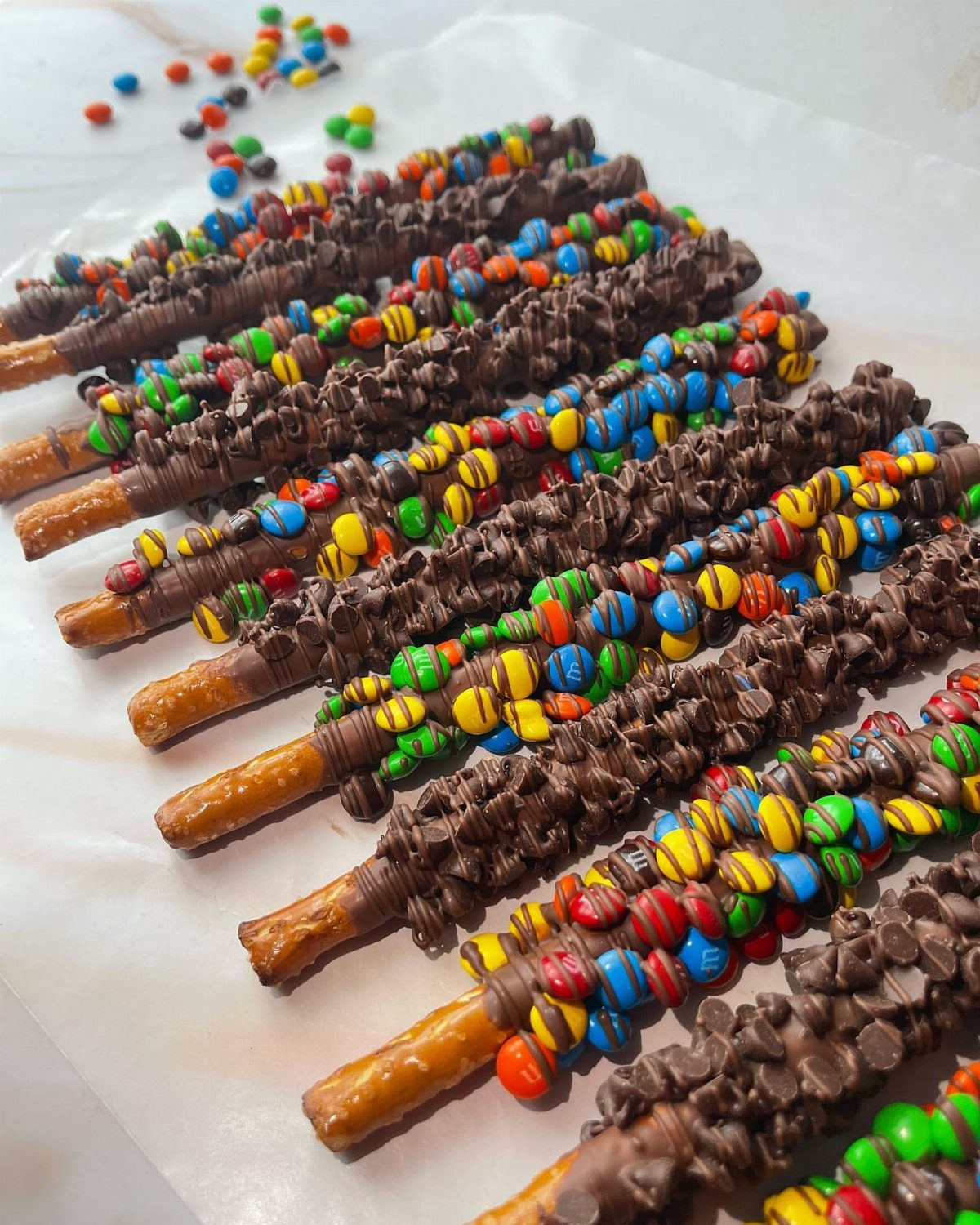 Chocolate-dipped pretzel rods decorated with colorful candy chocolates on white parchment paper.