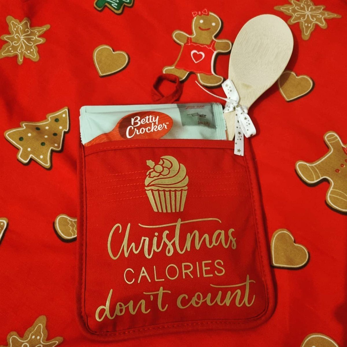 Red Christmas gift bag containing cookie mix and a wooden spoon among gingerbread cookies.