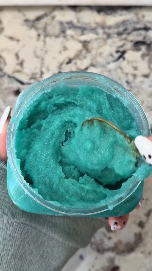 Turquoise sugar scrub in a glass jar being scooped with a small spoon.