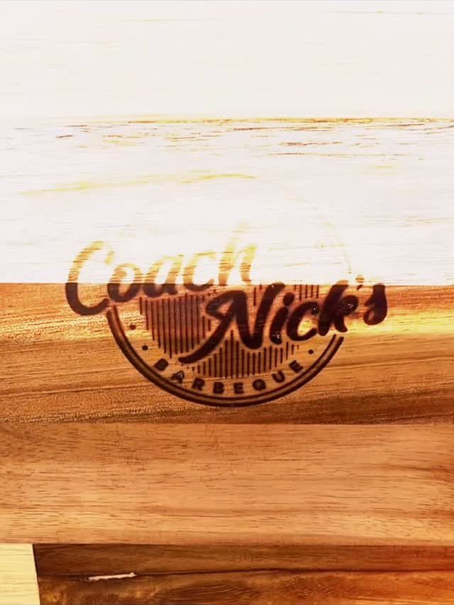 Wooden cutting board with an engraved Coach Nick's Barbeque logo on the surface.