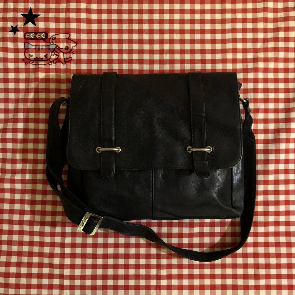Black leather messenger bag with silver buckles on a red checkered background