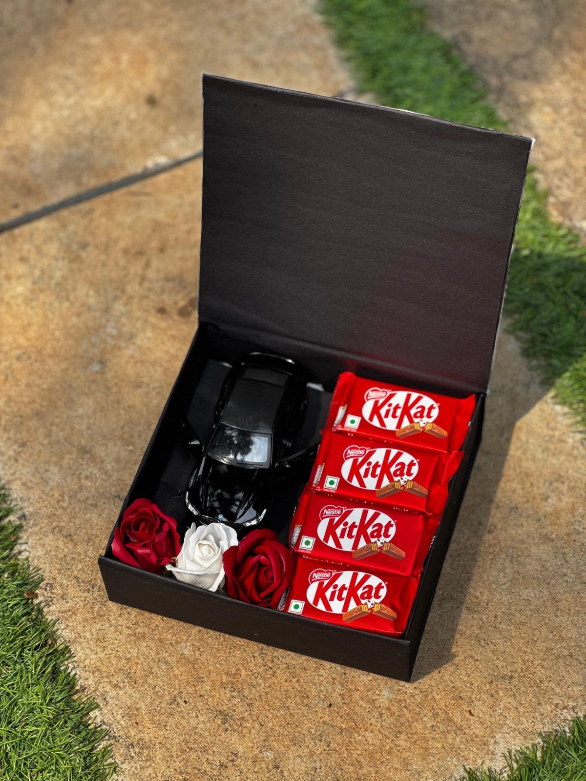 Black gift box with a toy car, KitKat chocolates, and artificial roses.