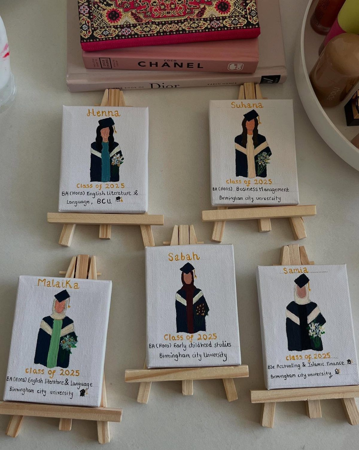 Five small illustrated graduation portraits on canvases resting on miniature wooden easels.