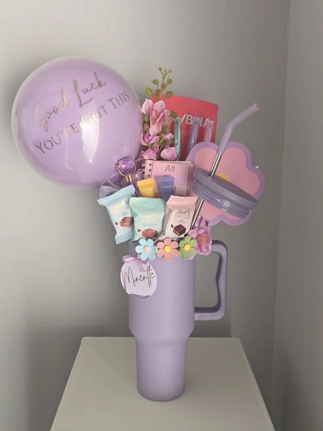Purple gift tumbler with snacks and a Good Luck balloon on white surface