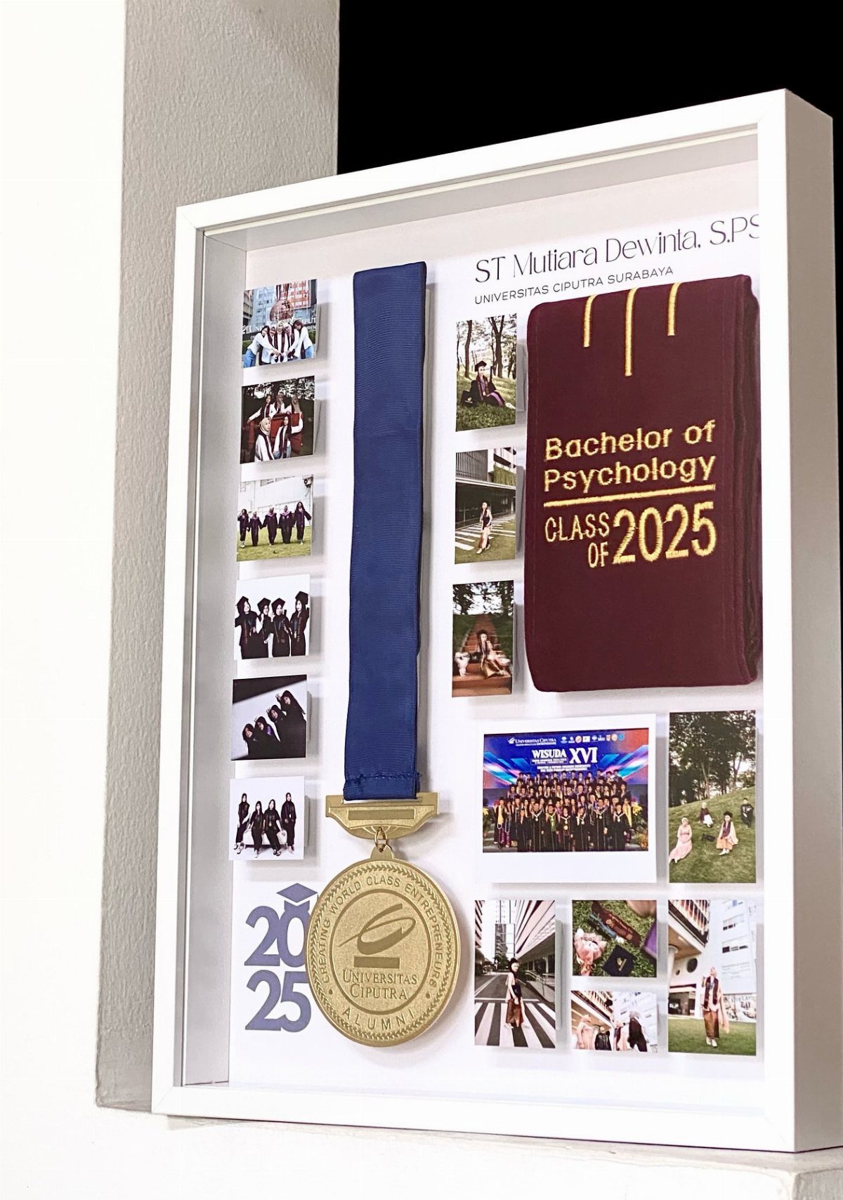 White shadow box frame with graduation diploma, gold medal, and small photos.