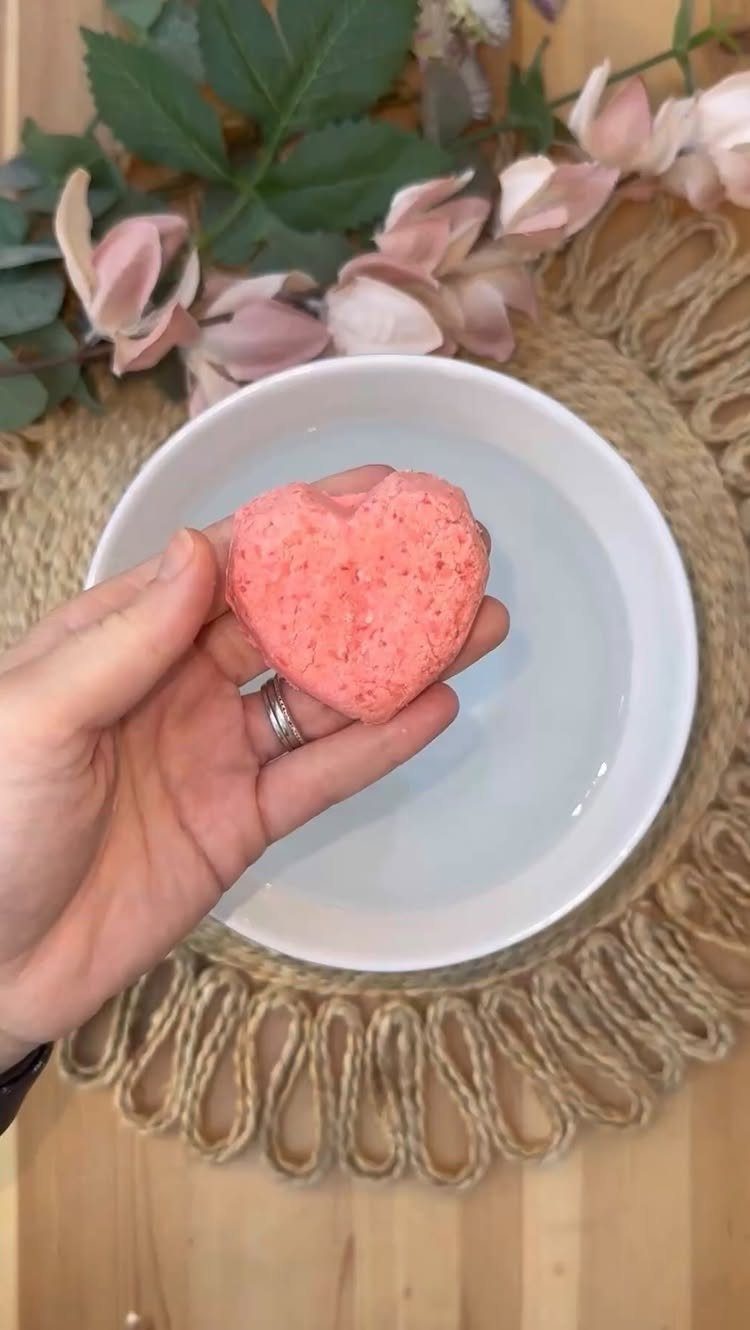 Pink heart-shaped bath bomb held over a white bowl of water with flowers.