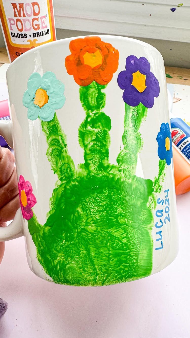 White ceramic mug decorated with a green handprint and colorful painted flowers.