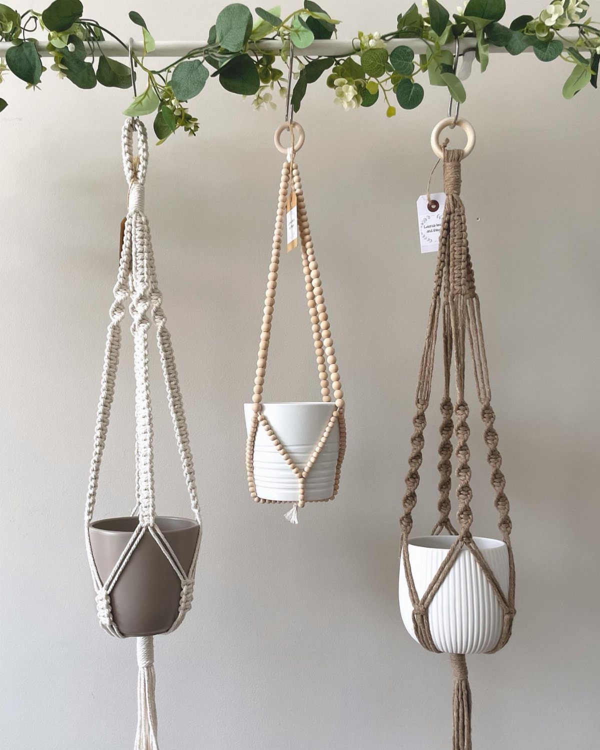 Three macrame plant hangers holding ceramic pots hanging from a leafy white rod.