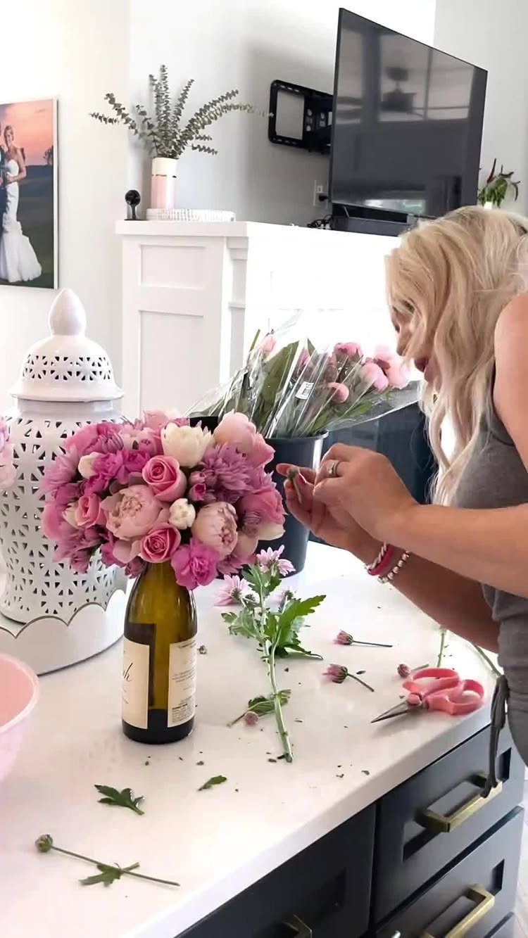 Woman arranging pink roses in a wine bottle vase on a white counter.