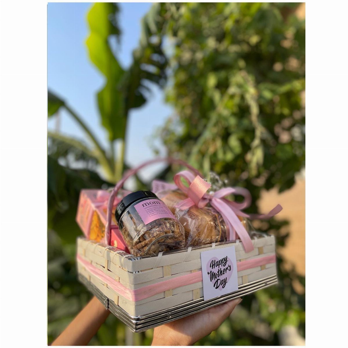 Gift basket with pink ribbons and treats held against a green garden background.
