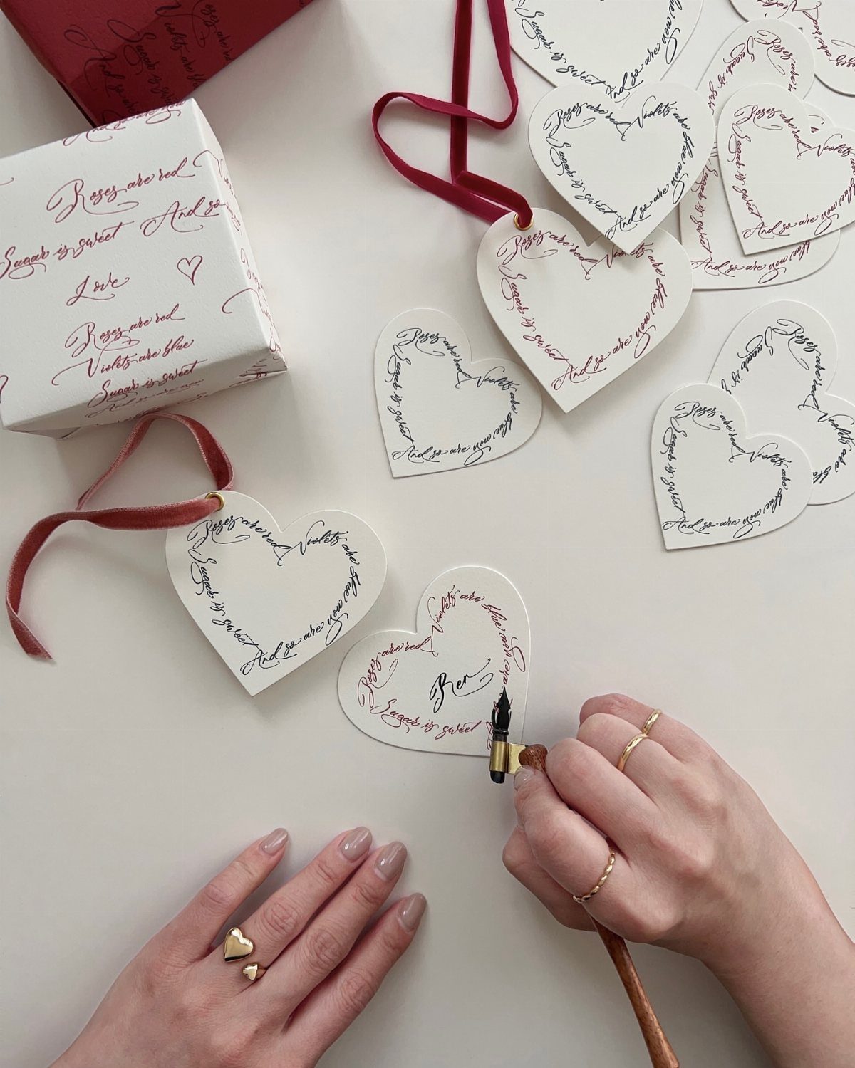 Hands writing calligraphy on heart-shaped white cardstock bookmarks with red ribbons.