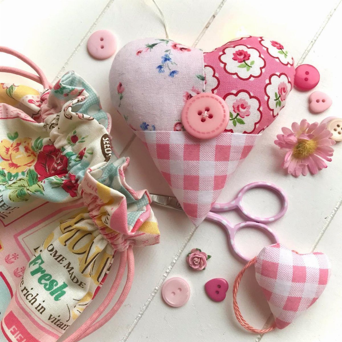 Heart-shaped fabric pincushion with floral and gingham patterns surrounded by sewing notions.