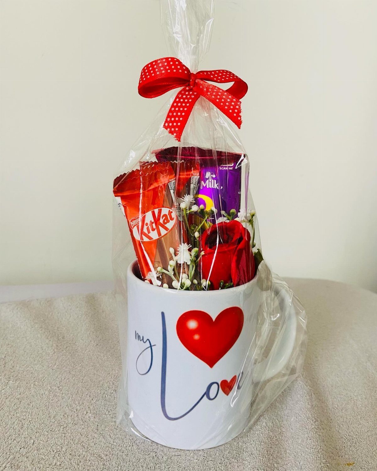 White mug with red heart filled with chocolates and a rose in cellophane.