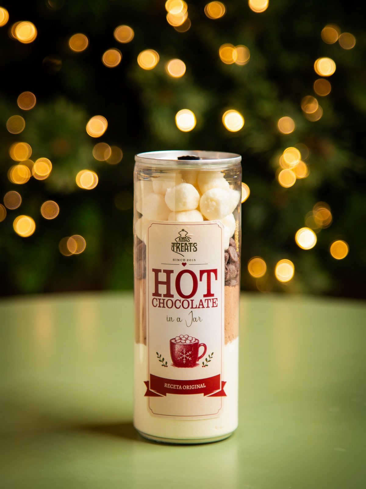 Glass jar filled with layered hot chocolate ingredients and marshmallows against festive lights.