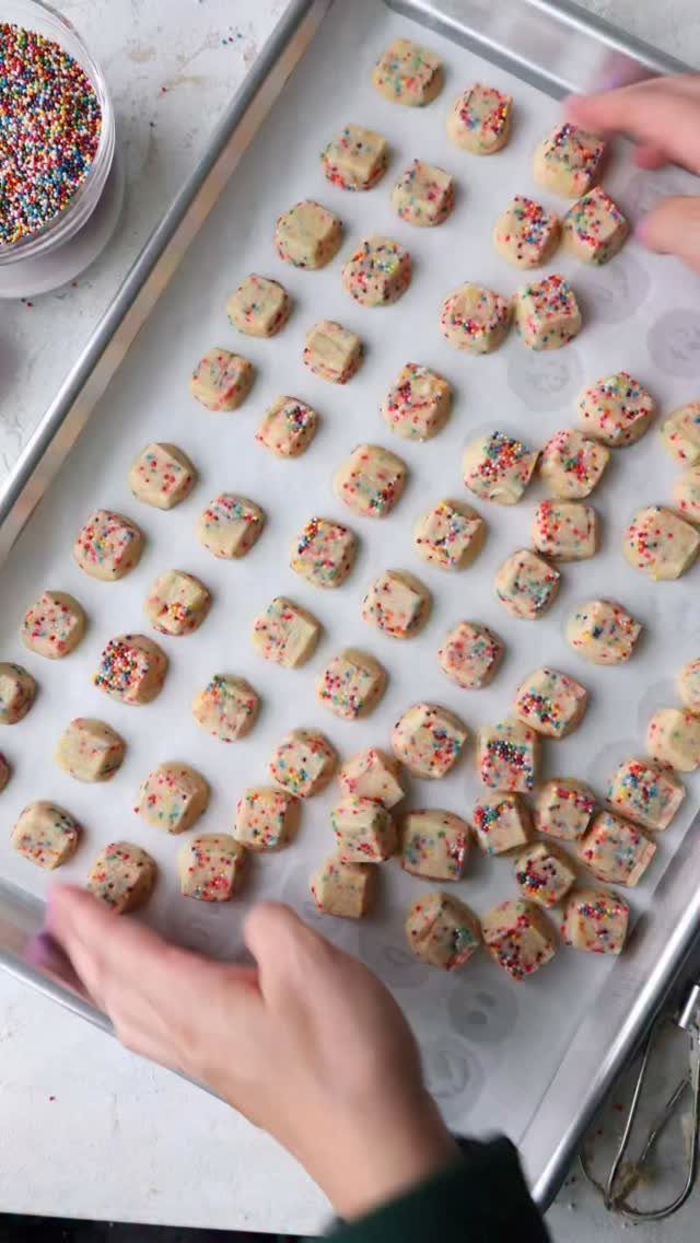 Small square shortbread cookies with rainbow sprinkles on a parchment-lined baking sheet.