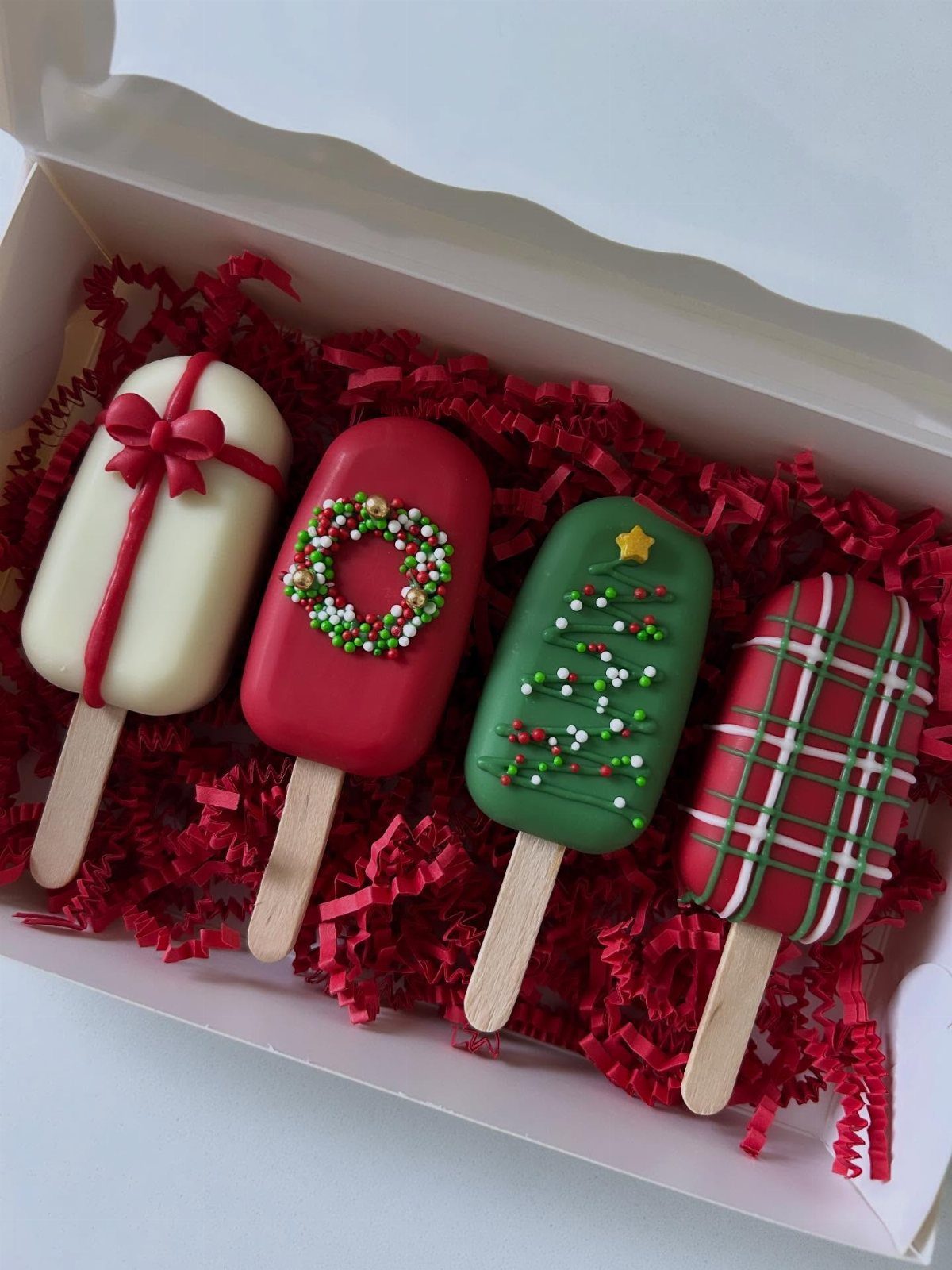 Christmas themed cake pops in a white box with red shredded paper filler.