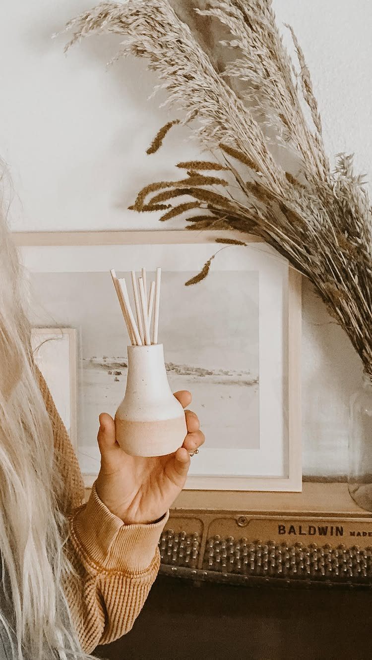 Hand holding a small ceramic reed diffuser with wooden sticks in a neutral room