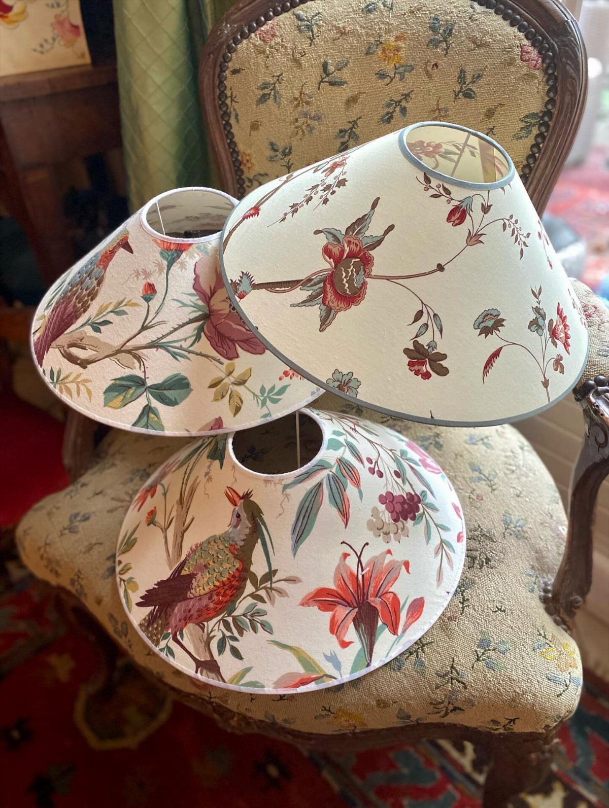 Three floral and bird patterned fabric lampshades resting on a vintage upholstered chair.