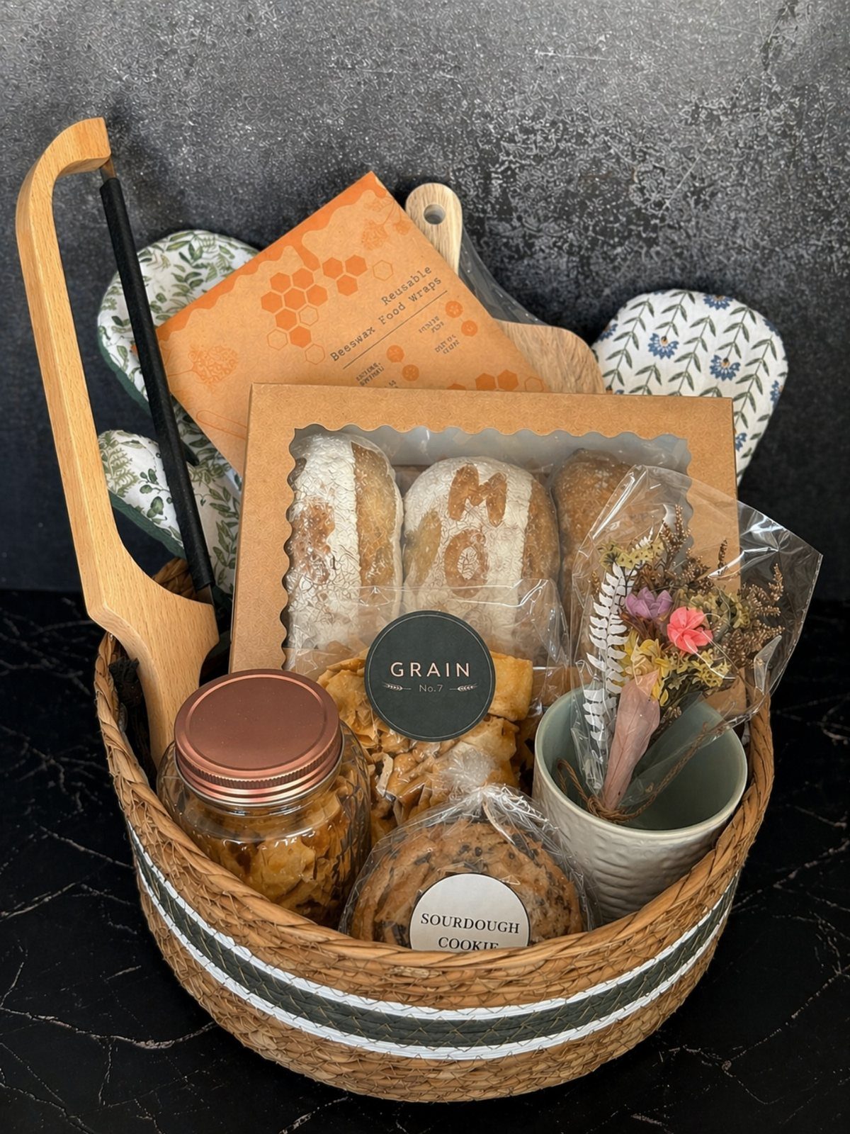 Woven gift basket containing fresh baked bread, a sourdough cookie, and kitchen accessories.