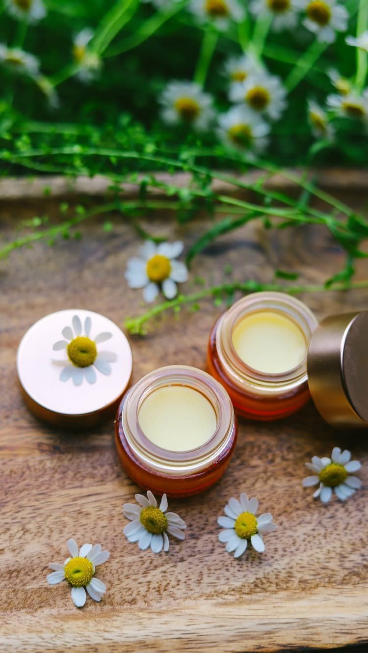 Two small jars of yellow balm with chamomile flowers on a wooden surface.