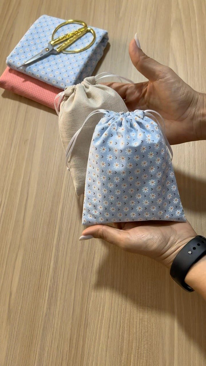 Two small drawstring cloth gift bags, one floral blue and one beige, held by hands.