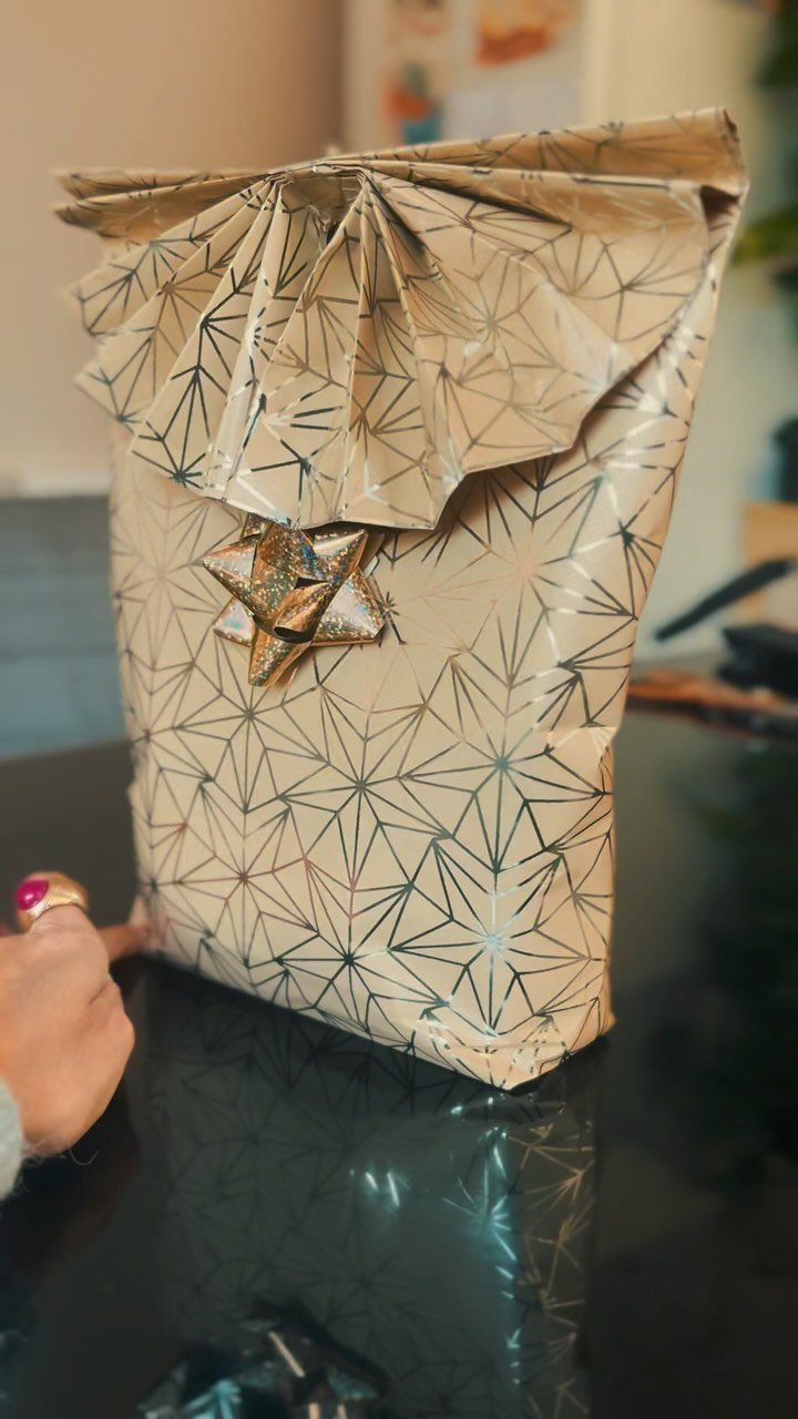 Wrapped gift with beige geometric paper, a fan-folded top, and a gold bow.