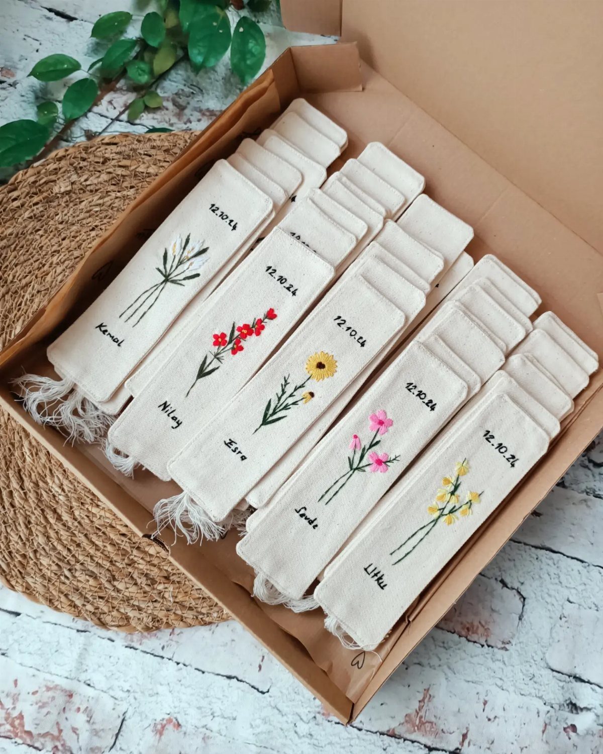 White fabric bookmarks with embroidered flowers and names arranged in a cardboard box.