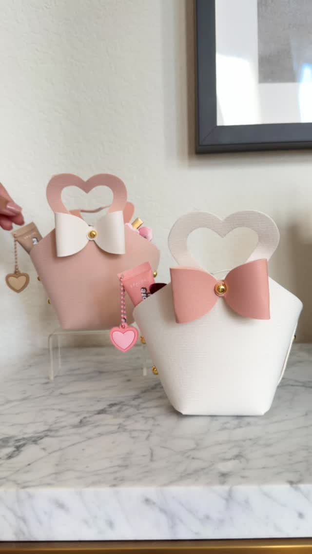 Two heart-shaped gift bags in pink and white with bows on marble surface.