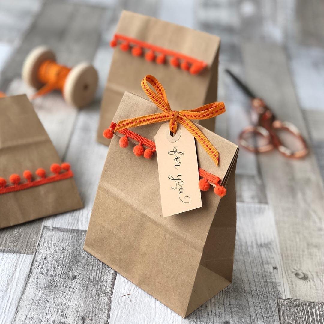 Brown kraft paper gift bags with orange pom-pom trim and ribbons on wood.