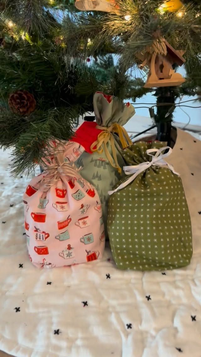 Three fabric drawstring gift bags in different patterns under a lit Christmas tree.