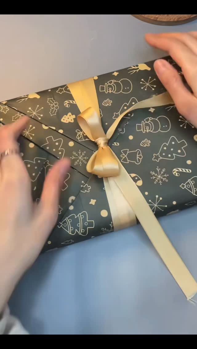 Hands tying a gold satin ribbon bow diagonally across a holiday gift box.