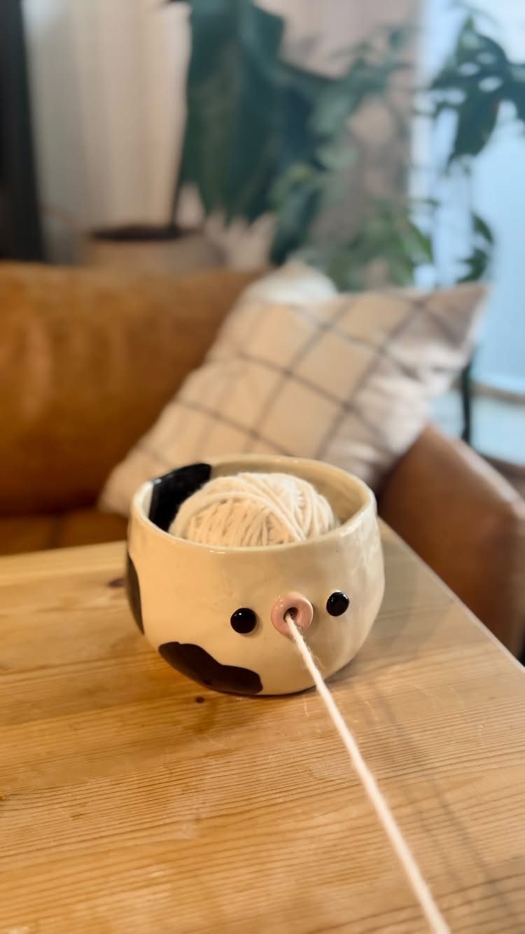 Cow-shaped ceramic yarn bowl on a wooden table holding white yarn