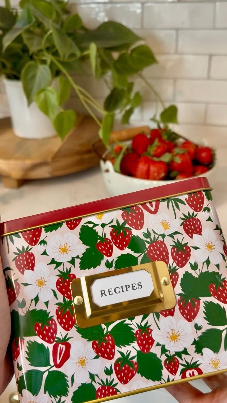 Strawberry patterned metal recipe box held in front of fresh strawberries and plants.