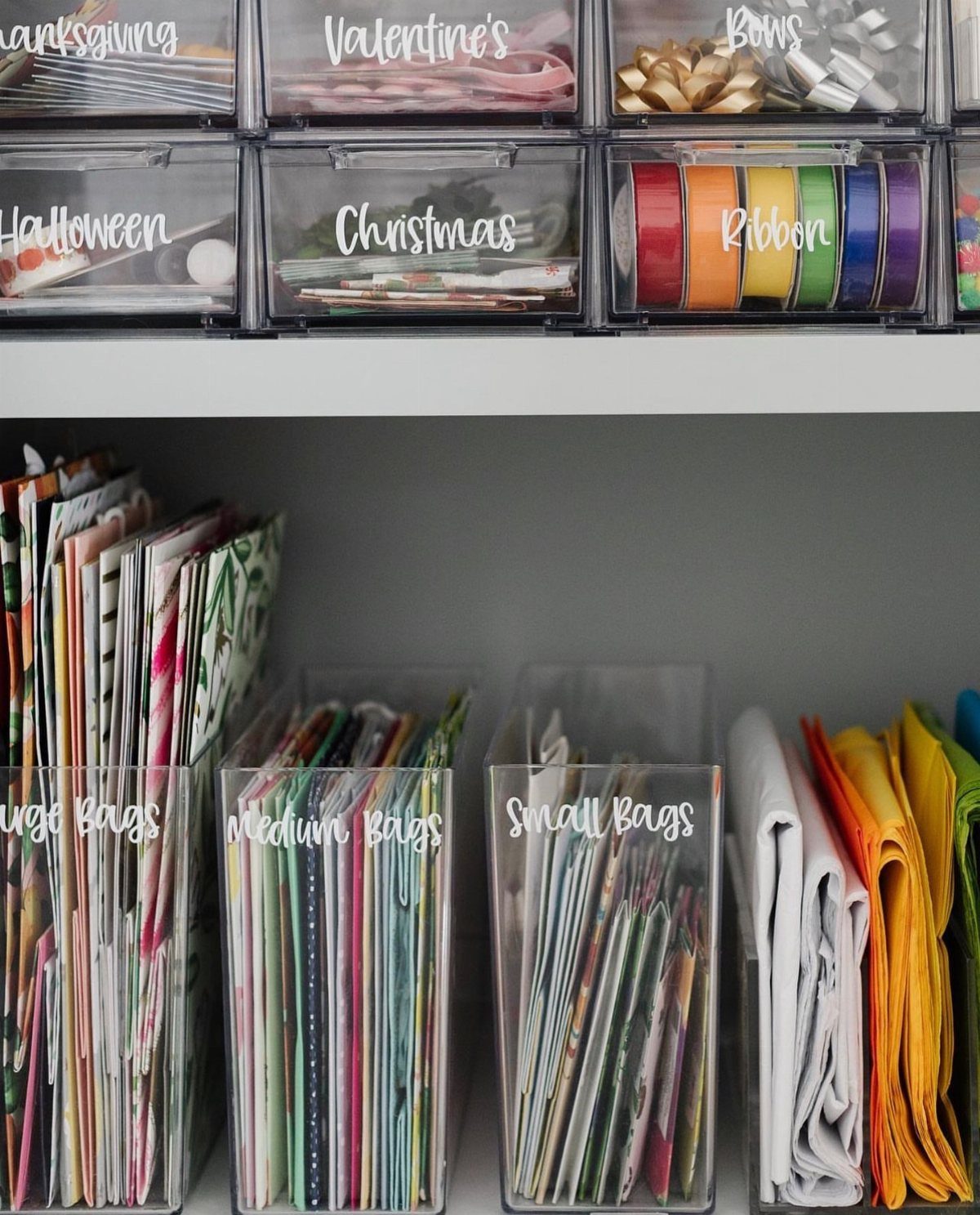 Organized gift wrap supplies in clear labeled plastic bins on white shelving units.