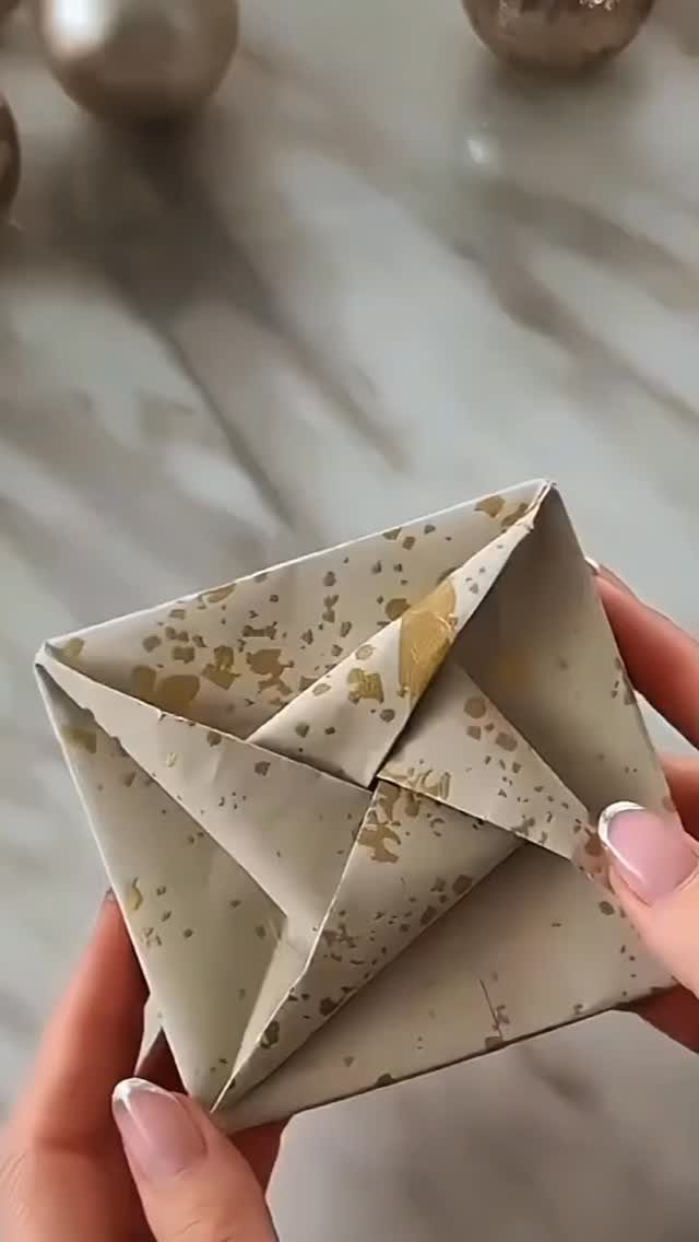 Hands holding a small gift wrapped in gold-speckled beige paper with origami folds.