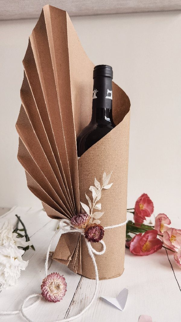 Wine bottle wrapped in brown paper with a pleated fan design and dried flowers.