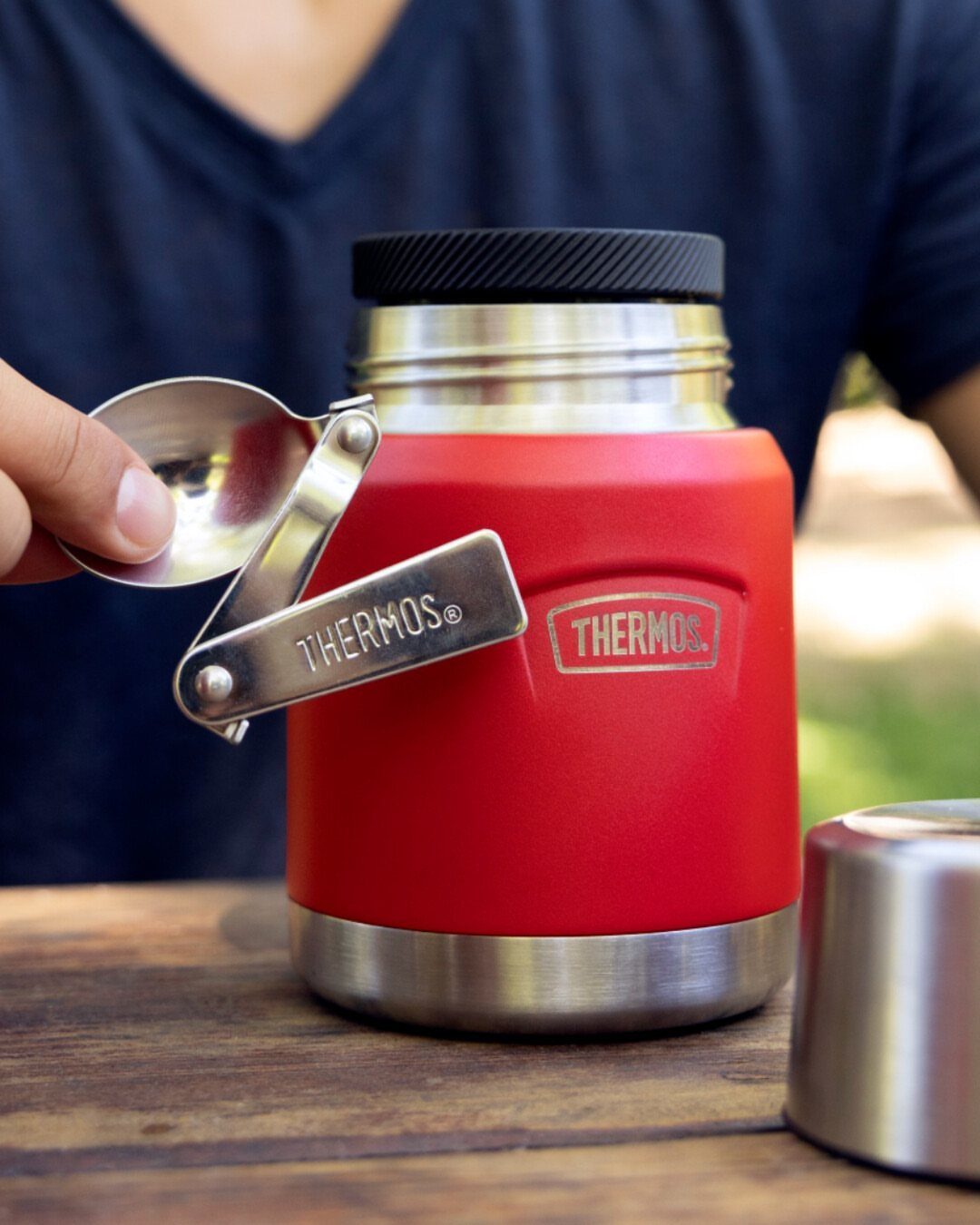 Red Thermos insulated food jar with a silver spoon on a wooden table.