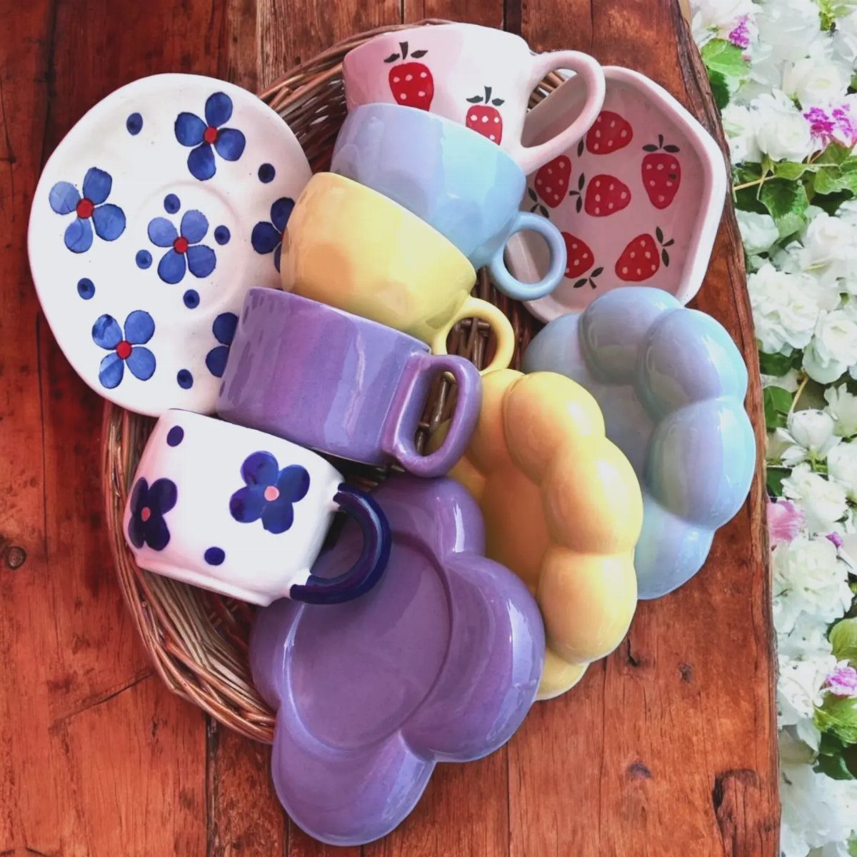 Colorful ceramic mugs and plates with floral and strawberry patterns in a wicker basket.