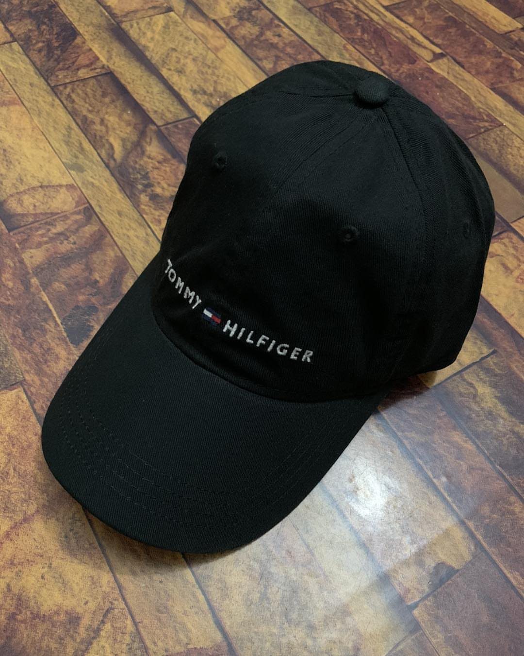 Black Tommy Hilfiger baseball cap with white embroidery on a wooden floor.