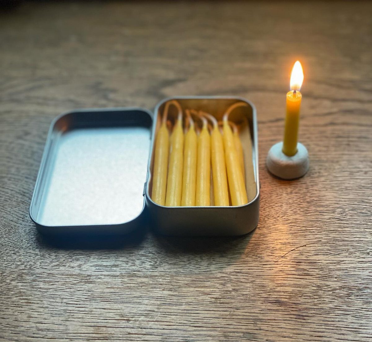 Yellow beeswax candles in a metal tin and one lit candle in stone holder.