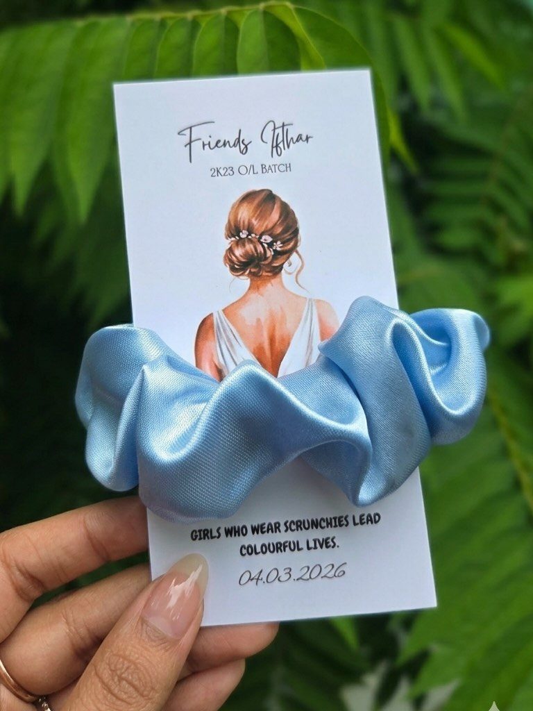Light blue satin scrunchie on a decorative white card held against green leaves.