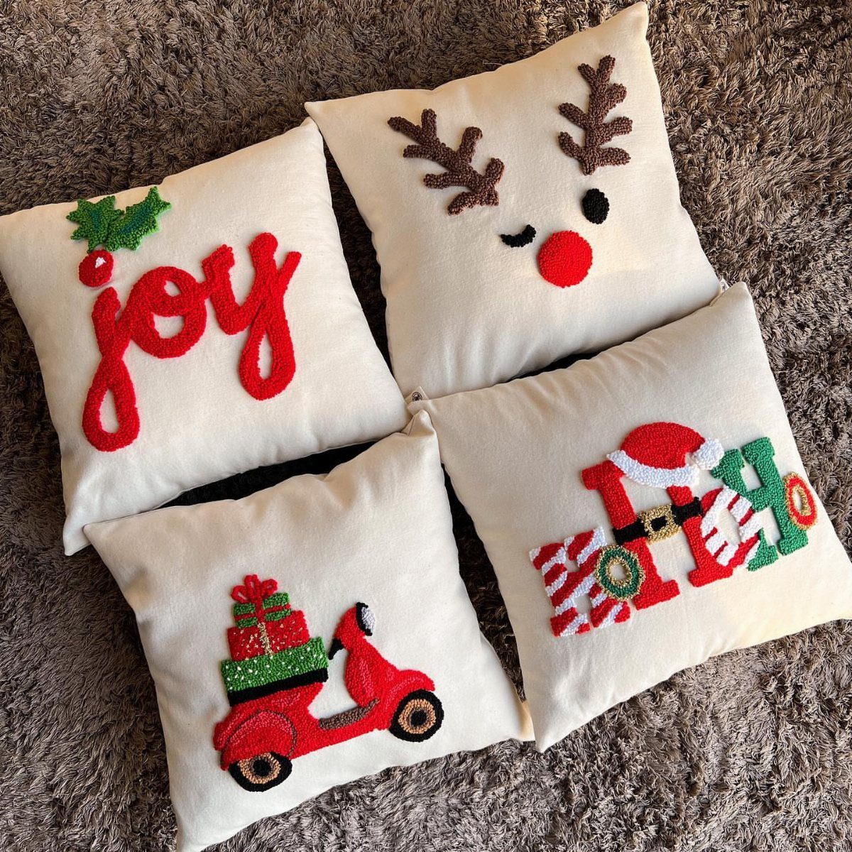 Four cream throw pillows with embroidered Christmas designs on a grey shag rug.