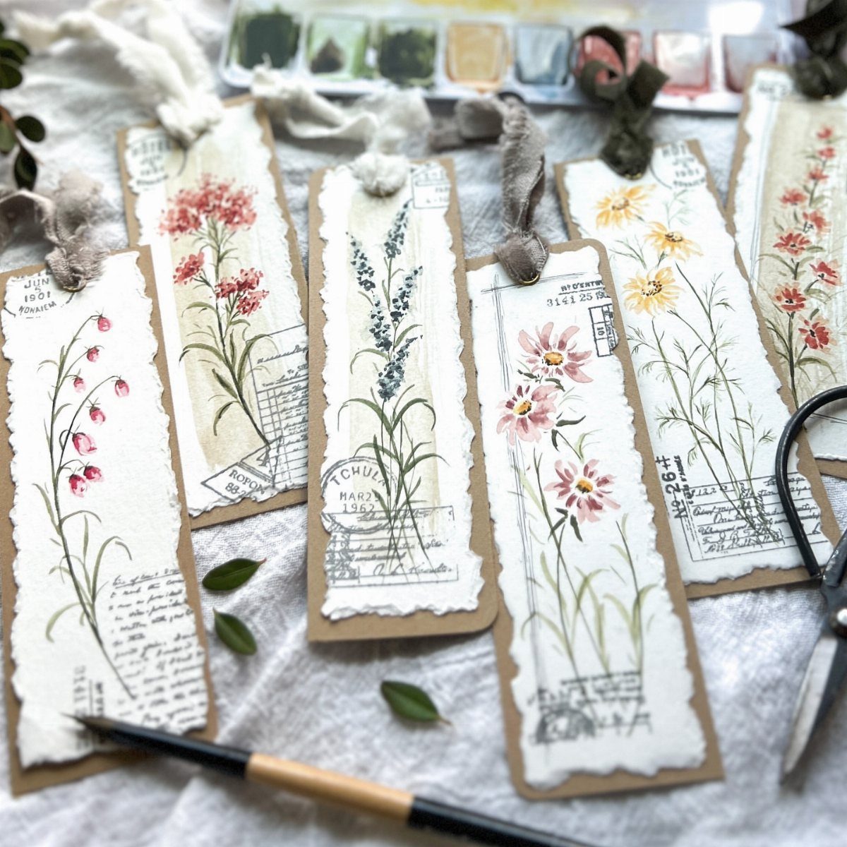 Handmade watercolor floral bookmarks with torn edges on a white fabric background.