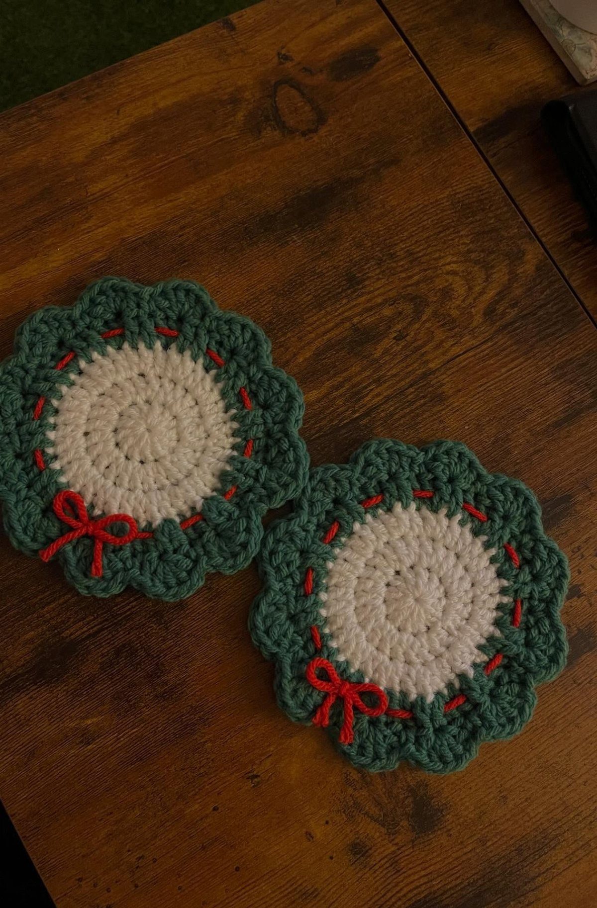 Two crocheted wreath coasters with green borders and red bows on wooden table.