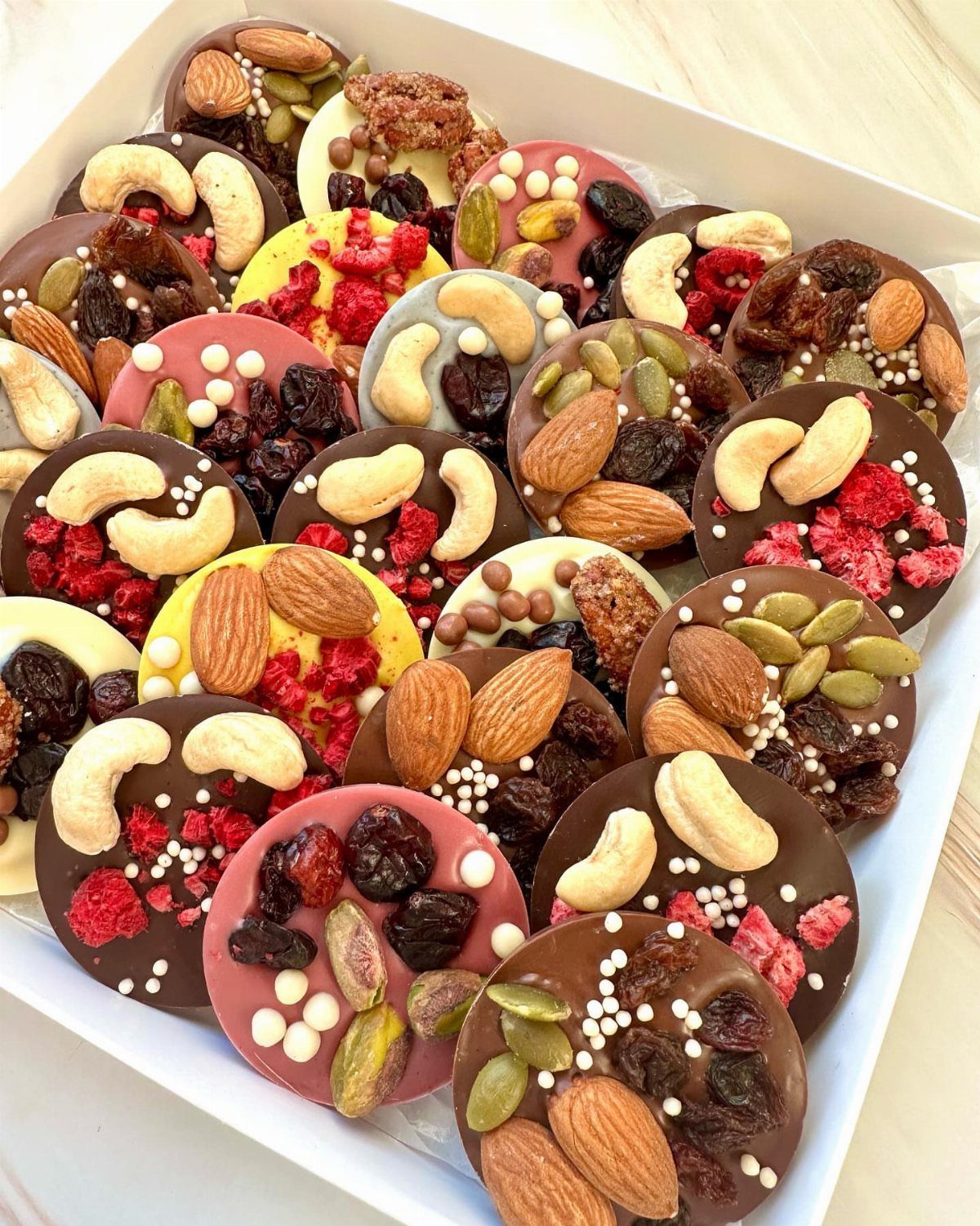 Round chocolate discs topped with assorted nuts and dried fruits in a white box.