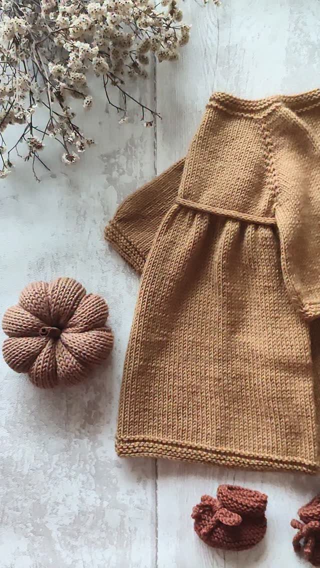 Brown knitted baby dress, booties, and a small pumpkin on a wooden surface.